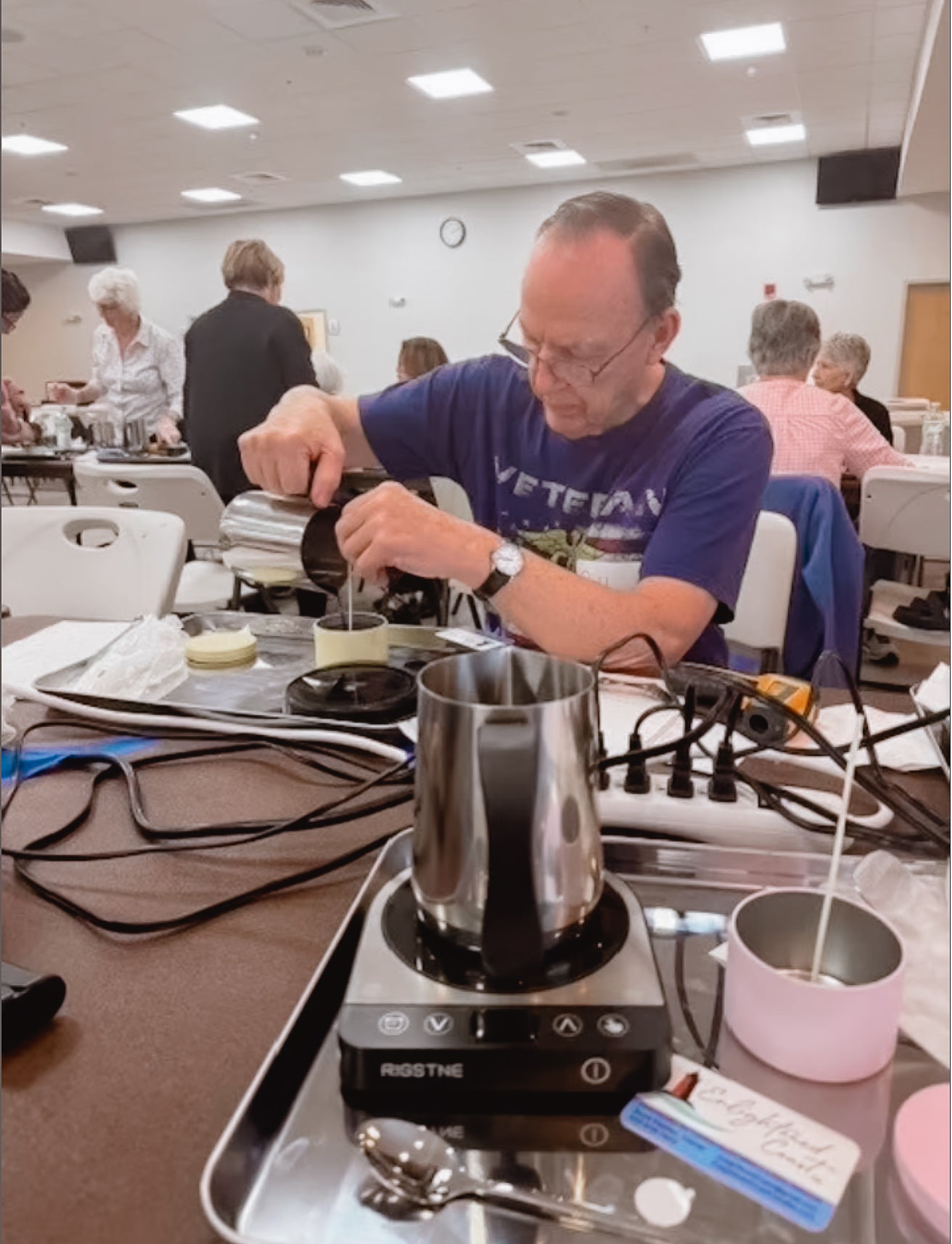 Person stirring a pot on a portable stove in a candle making workshop setting with other people in the background.