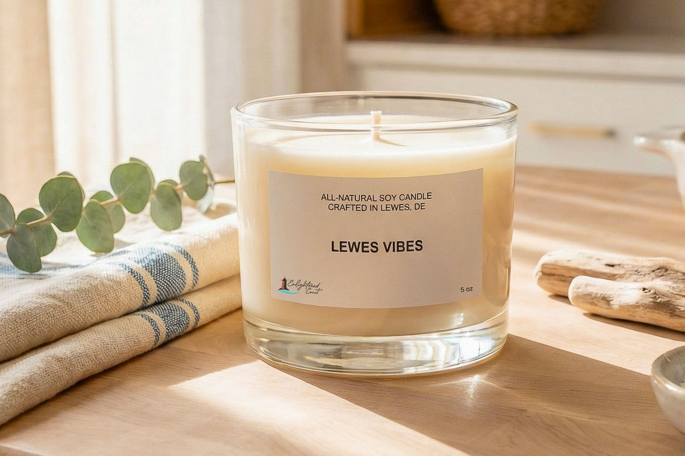 Candle labeled 'LEWES VIBES' on a wooden surface with eucalyptus leaves and folded towels in the background.