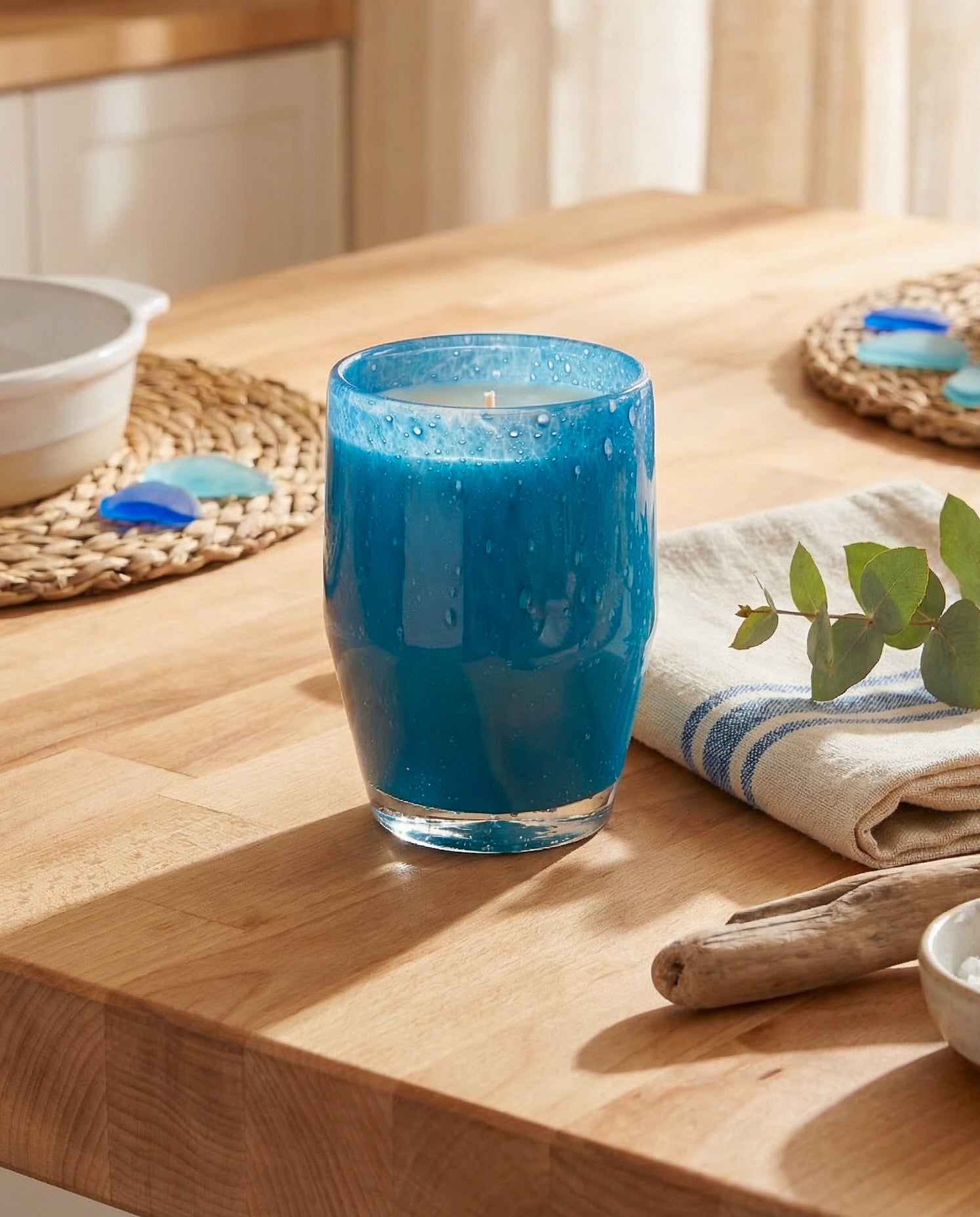 Teal candle in a glass container on a wooden surface with a plant and towel in the background