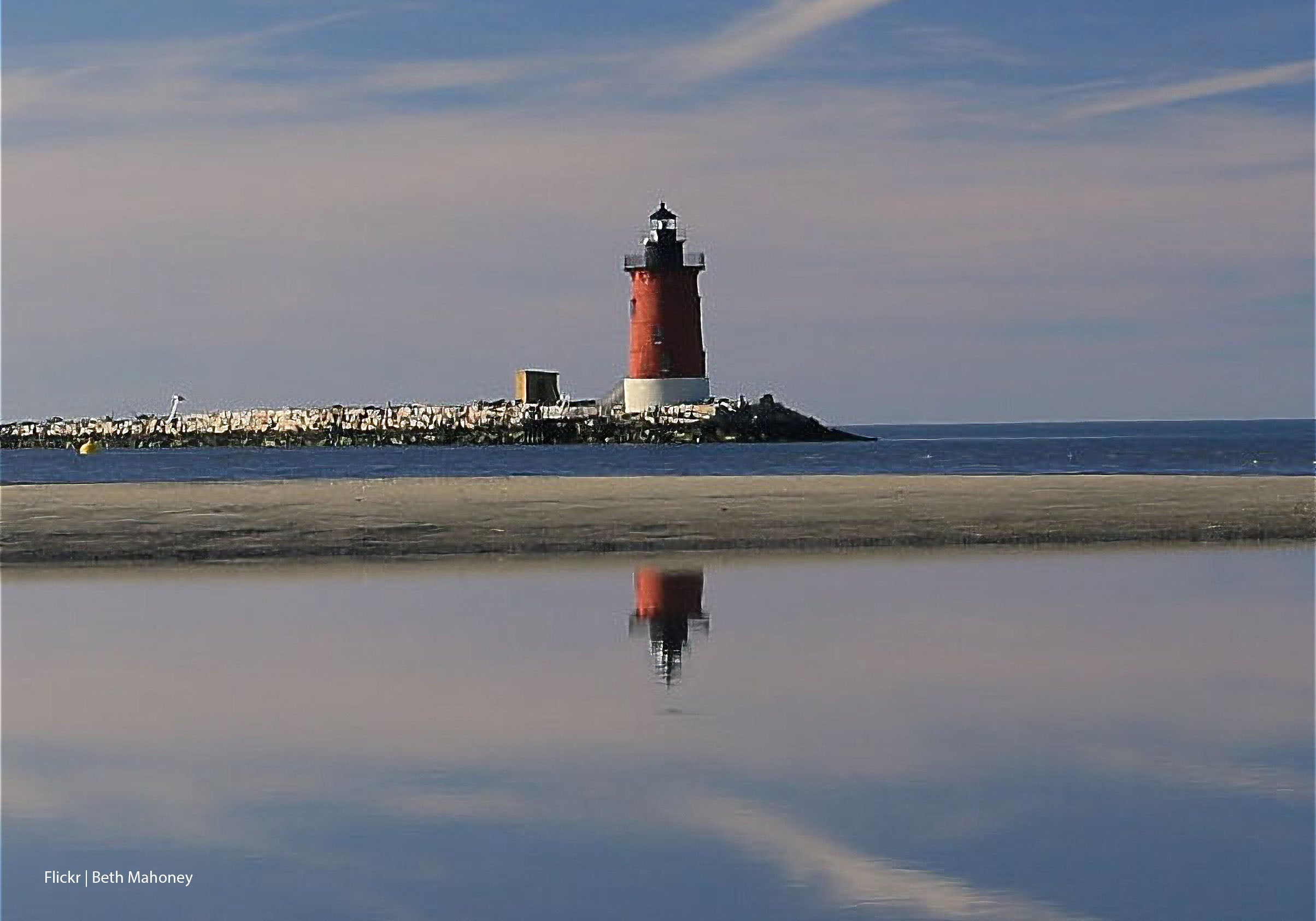 Red and white Lewes lighthouse reflected in calm water with a clear sky.